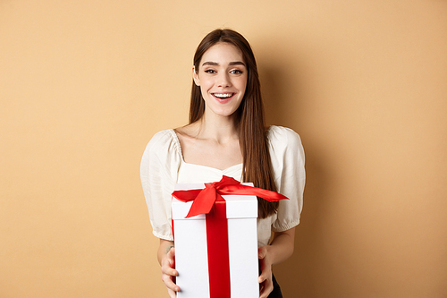 Happy valentines day. Cheerful smiling girl giving surprise gift to lover,  and holding present in box, standing on beige background.