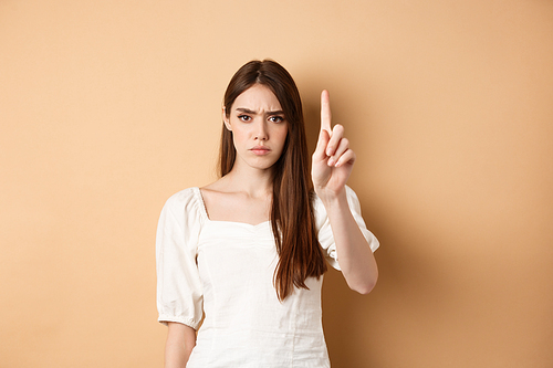 Rule number one. Serious frowning woman showing finger to scold person, look disappointed and angry, standing on beige background.