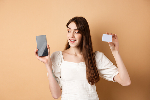 Check this out. Smiling happy young woman showing plastic credit card and empty mobile phone screen, demonstrate application, standing on beige background.