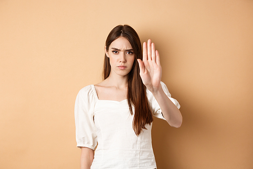 Nope stop right there. Serious and confident woman stretch out hand to forbid something, frowning and saying no, disagree and reject bad offer, standing on beige background.