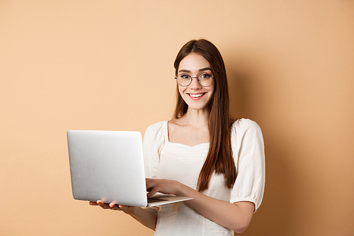 Smiling girl student working on laptop, wearing glasses and looking happy, using computer while standing against beige background.
