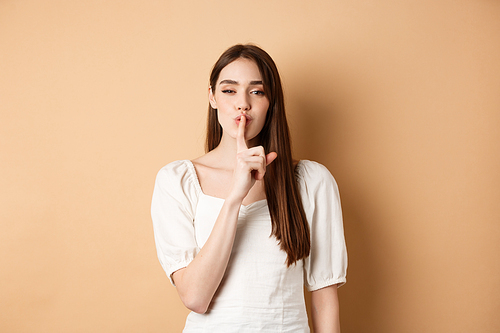 Sensual cute girl hiding secret, showing hush gesture, make shhh sound with finger on lips, stay quiet sign, standing on beige background.