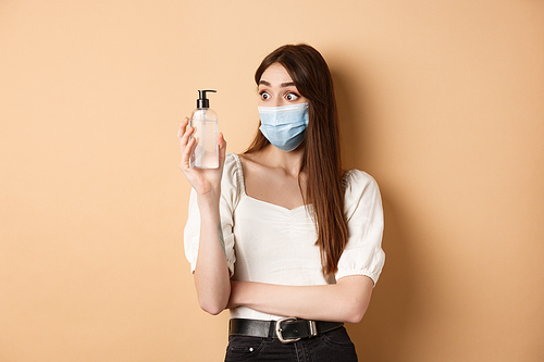 Covid-19 and preventive measures concept. Excited girl in medical mask look amazed at hand sanitizer bottle, standing on beige background.