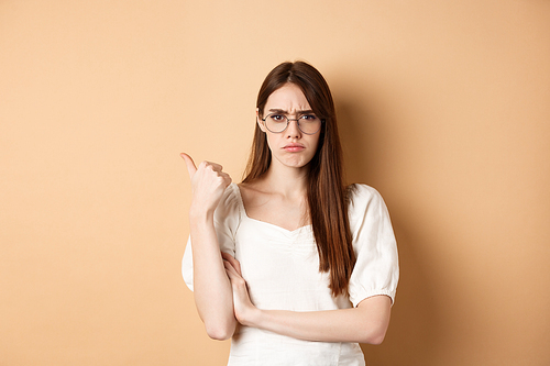 Disappointed girl in glasses frowning, pointing aside at bad product, disapprove and dislike something, standing on beige background.