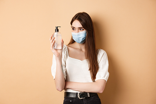 Covid-19 and preventive measures concept. Smiling girl in face mask look at hand sanitizer bottle, using hygiene product from coronavirus outbreak, beige background.