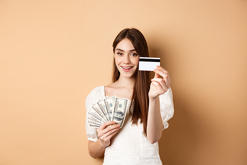 Smiling girl showing platic credit card to camera, holding dollar bills close to chest, standing on beige background.