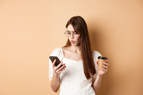 Shocked young woman reading mobile phone screen and frowning, holding coffee cup, standing on beige background.