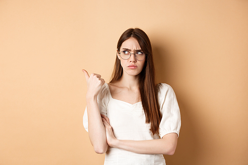 Suspicious woman in glasses pointing and looking aside, frowning with doubtful face, standing on beige background.
