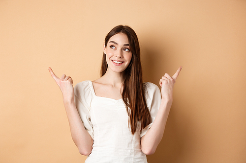Dreamy caucasian girl making choice, pointing fingers sideways and looking at upper left corner pensive, picking product, standing on beige background.