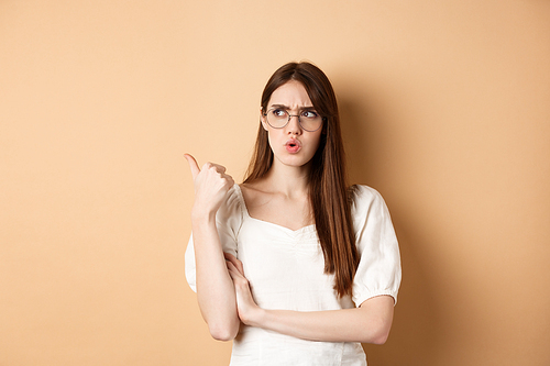 Confused and suspicious young woman in glasses frowning, pointing and looking aside with displeased face expression, standing on beige background.