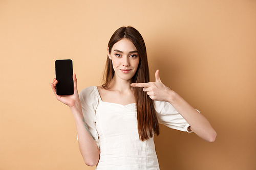 Confident girl pointing at empty smartphone screen, showing online promo deal, standing on beige background.