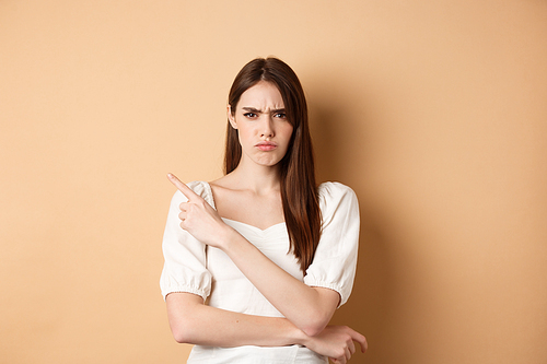 Angry frowning girl pointing left at empty space, looking disappointed and upset, scolding you or accuse, standing on beige background.