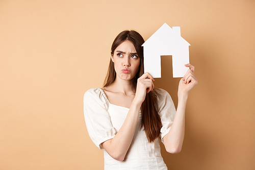 Real estate. Upset young woman showing paper house cutout and frowning sad, looking aside thoughtful, thinking of buying apartment, standing on beige background.