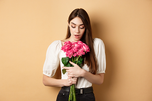 Valentines day. Surprised tender girl looking at beautiful pink roses, receive romantic gift from lover, standing on beige background.