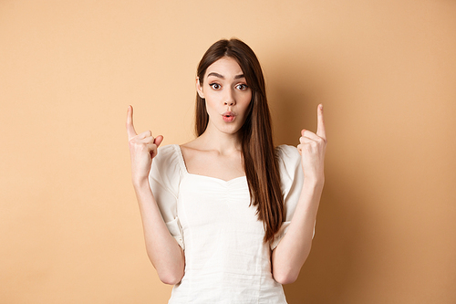 Wow look. Intrigued young woman showing interesting offer, pointing fingers up and look excited, standing on beige background.