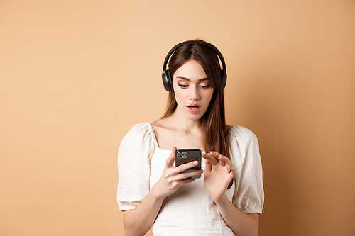 Excited girl listen music in headphones and looking at smartphone amazed, reading awesome news, found cool playlist, standing on beige background.