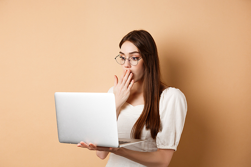 Shocked girl look at laptop screen, covering mouth with amazed face, standing in glasses on beige background.