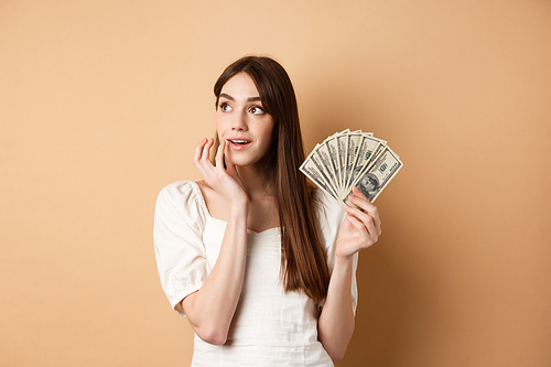 Dreamy girl holding dollar bills and thinking off shopping, looking at upper left corner thoughtful, plan how to waste money, standing on beige background.