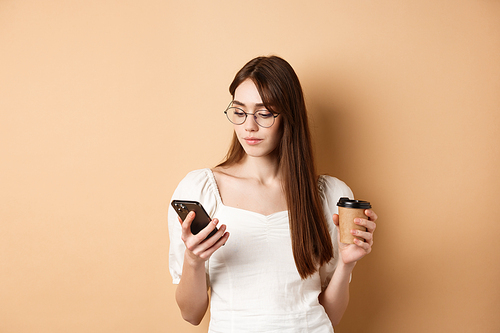busy girl in glasses reading message on mobile phone and  coffee takeout, standing on beige background.