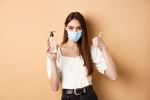 Covid-19 and preventive measures concept. Smiling girl in medical mask show thumb up and hand sanitizer, recommend product for disinfection, beige background.
