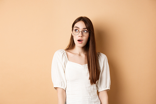 Excited young woman in glasses looking aside with opened mouth, gasping fascinated, standing on beige background.