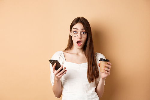Surprised girl react to mobile phone news, holding smartphone and cup of coffee, look excited with dropped jaw at camera, beige background.
