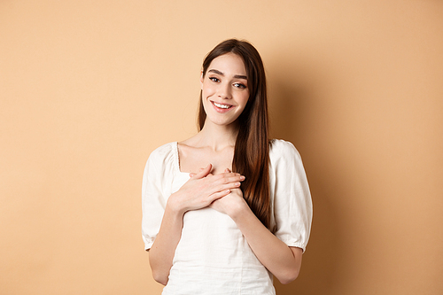 Pretty girl holding hands on heart and smiling, thanking you, feeling grateful and pleased, standing in dress on beige background.