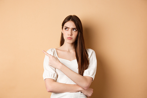 Suspicious woman in dress looking, pointing left, frowning and staring with disbelief and doubts, standing on beige background.