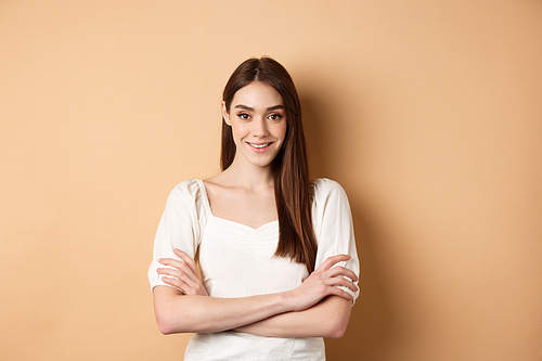 Smiling candid girl in dress cross arms on chest, looking like professional, standing confident on beige background.