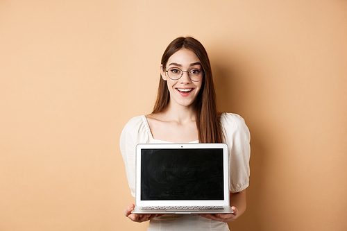 Happy woman in glasses showing laptop screen and smiling, demonstrate internet promo deal, standing on beige background.