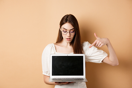 Curious girl in glasses pointing at laptop screen, check out online deal, demonstrate project on computer, standing on beige background.