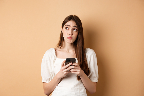 Confused young woman thinking after reading news on mobile phone, looking at upper left corner hesitant, standing on beige background.