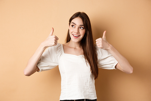 Smiling dreamy girl looking at upper left corner and showing thumbs up at promo, recommending product, praising banner, standing on beige background.