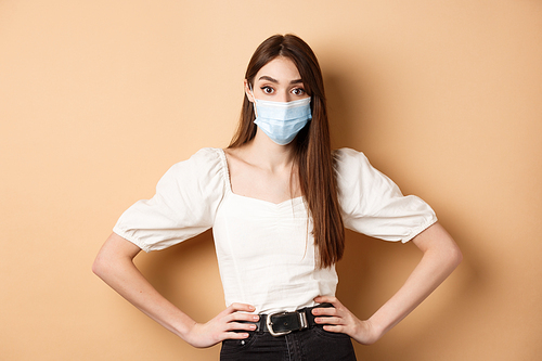 Covid-19 and lifestyle concept. Woman in medical mask looking alarmed at camera, standing on beige background.