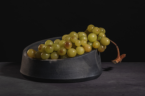 Modern design black ceramic bowl with vine of green grapes on dark countertop and background.