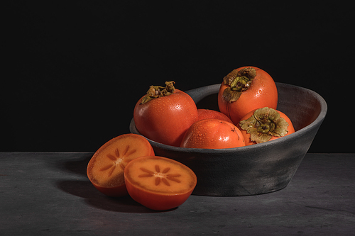 Modern design black ceramic bowl with persimmon fruits  on dark countertop and background.