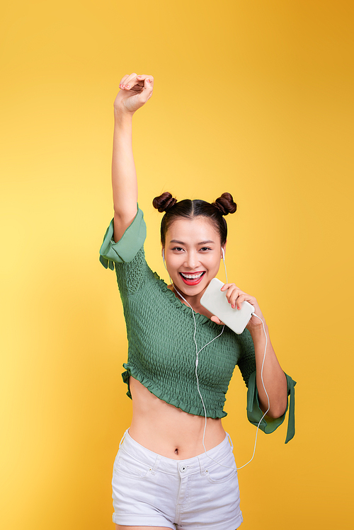 Fashion smiling asian woman listening to music in earphones  over yellow background