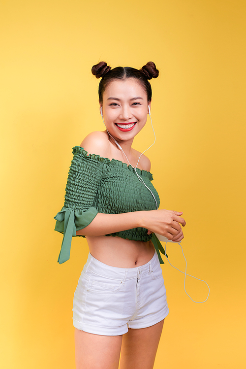 Fashion smiling asian woman listening to music in earphones  over yellow background