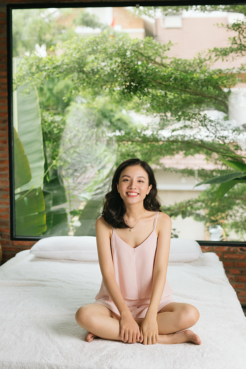 A woman is sitting on her bed with her legs folded, her arms resting on her legs and smiling
