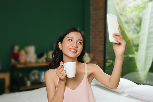 Young woman sitting on bed with cup of coffee selfie on mobile phone