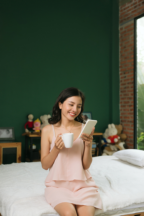 Relaxed young woman sitting on bed with a cup of coffee and cellphone