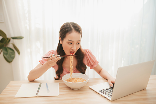 Portrait of young Asian woman freelancer working with laptop at her home and eating instant noodle