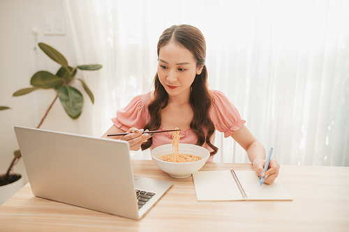 Girl eating instant noodle in hurry time while running her business at home on notebook computer
