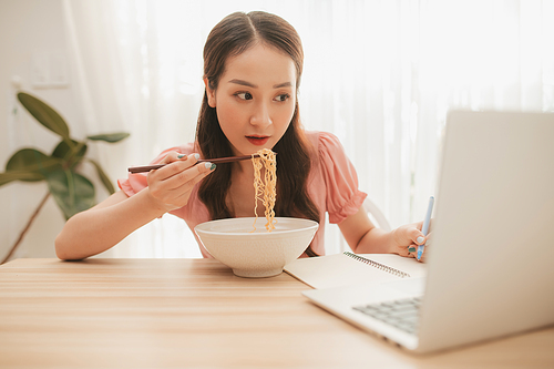Female employee holding chopsticks eating instant noodle at workplace in home office