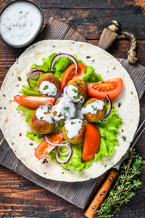 Vegetarian falafel with vegetables and tzatziki sauce on a tortilla bread. Dark wooden background. Top view.