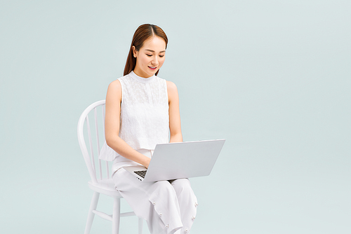 Full length of a happy young woman casually dressed sitting on a chair isolated over white background, working on laptop computer