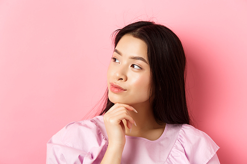 Close-up portrait of romantic asian girl looking aside at logo, thinking or daydreaming, standing against pink background.