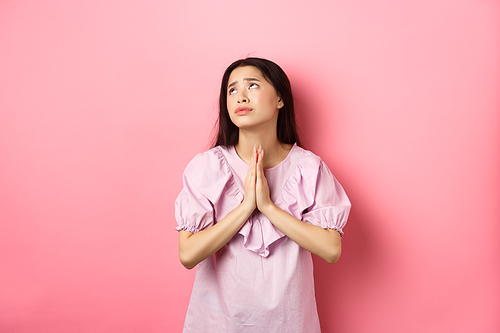 Hopeful asian girl praying god, looking up to plead or pray, making wish, supplicating with dramatic face, standing against pink background.