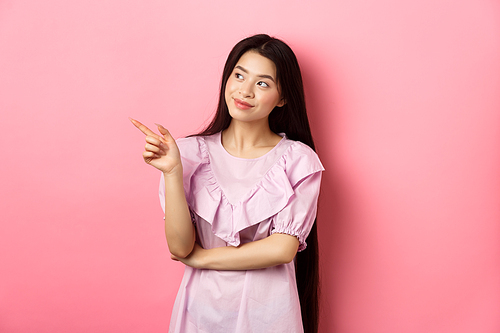 Dreamy asian girl in dress, looking and pointing left at logo, gazing at planner, standing against pink background.