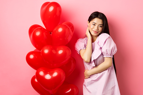 Beautiful asian girl in dress smiling coquettish, flirting on valentines day, looking sensual at camera, posing near valentines heart balloons, pink background.